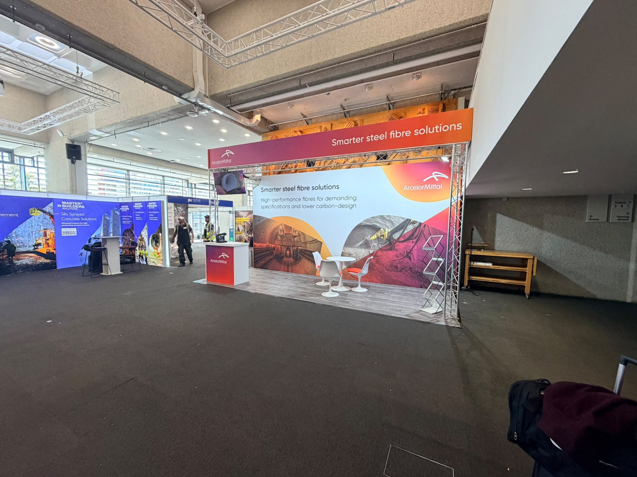 Full-height printed graphic wall on the ArcelorMittal stand showing tunnelling and sprayed concrete imagery, with white tulip seating on a timber-effect raised floor