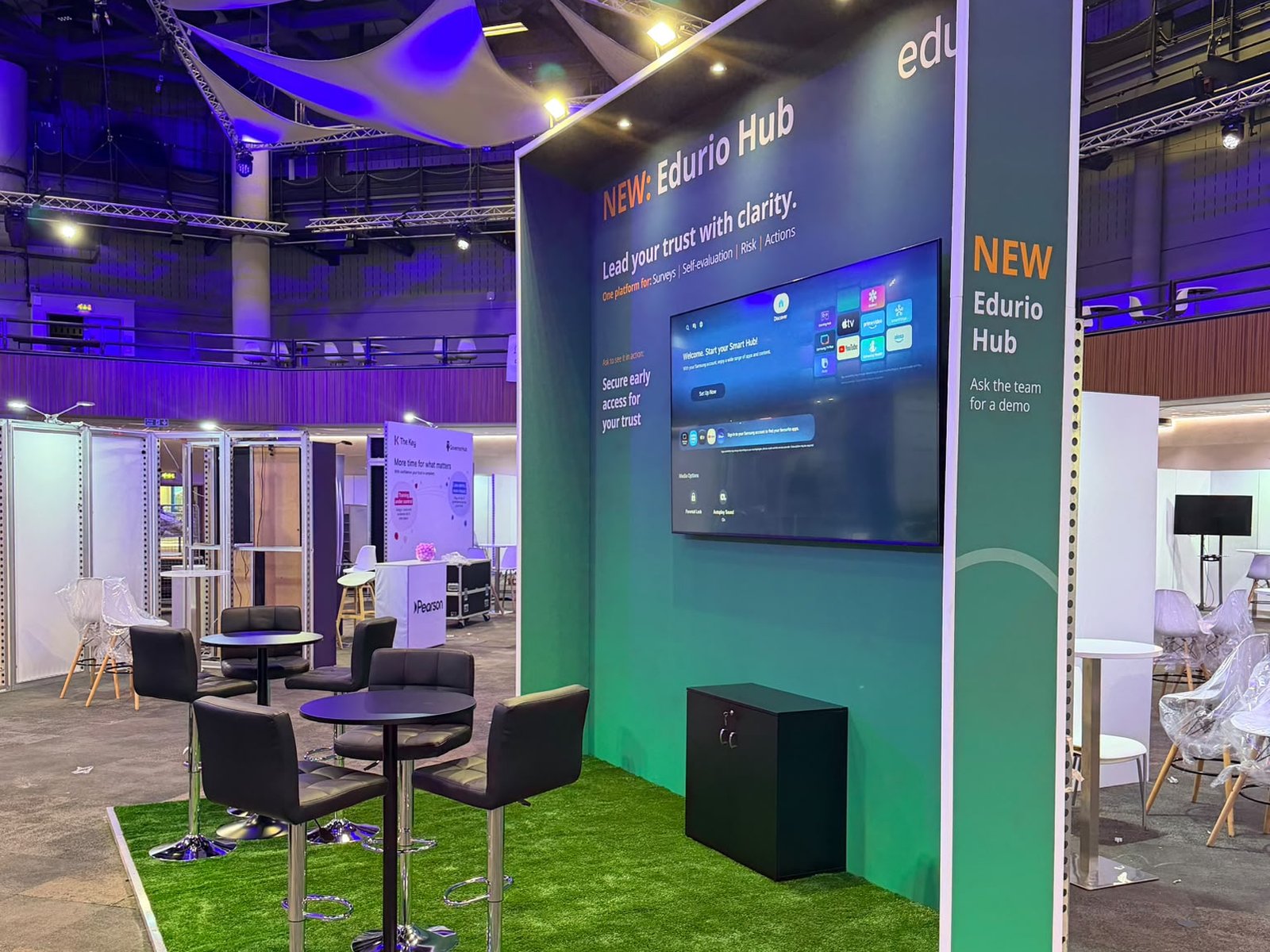 Side view of the Edurio stand at the ICC Birmingham, showing the bookend panel 'NEW Edurio Hub, Ask the team for a demo' and the canopy lights